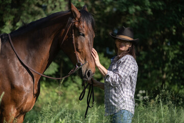 Fototapeta premium Woman wearing a hat rides a brown horse through tall grass in a vast open field under a clear blue sky with distant trees on the horizon