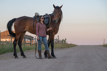 Woman wearing a hat rides a brown horse through tall grass in a vast open field under a clear blue sky with distant trees on the horizon