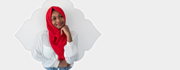 Naklejka premium A woman wearing a red hijab is sitting indoors. She has a thoughtful expression as she rests her chin on her hand. The background is light and simple, adding focus to her.