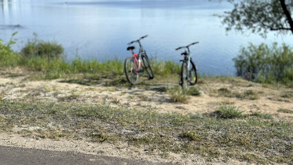 Bicycles on sandy riverbank in summer, resting in grassy park near calm river, sunlit shoreline with visible handlebars and tire