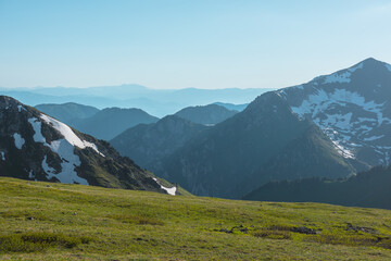 Fototapeta premium Scenic view from sunlit grassy meadow to abyss of alpine valley between big rocky mountains with snows and hilly layers of ridge silhouettes far away under clear blue sky in bright sun in sunny day.