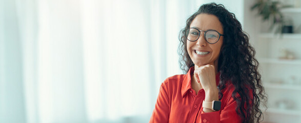 A woman with curly hair smiles while resting her chin on her hand. She is wearing glasses and a red...