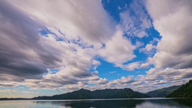 Time lapse stunning landscape view of Khun Dan Prakan Chon Dam showcasing calm blue waters reflecting dramatic white clouds and lush green mountains, creating a peaceful natural panorama.
