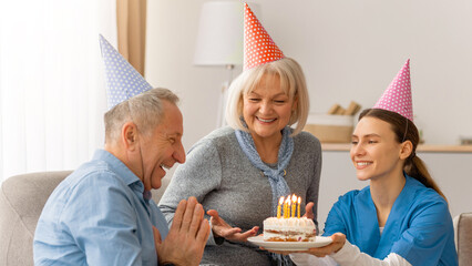 Obraz premium A man smiles and claps as he receives a birthday cake from a woman wearing blue scrubs. Another woman with gray hair smiles beside him. They celebrate together in a warm living room.