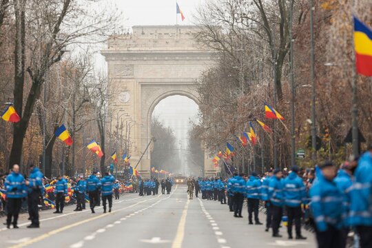 Festive atmosphere fills the streets of Bucharest as people gather near the Arc de Triumf to celebrate a national holiday. Bright flags wave in the air, creating a tapestry of colors