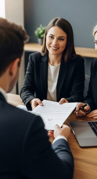 Smiling businesswoman in black suit handing over contract document to male client during a professional business meeting or interview, showcasing agreement and partnership