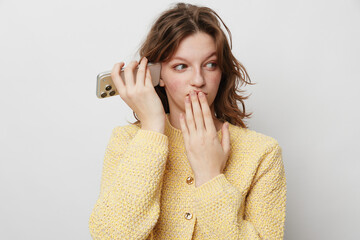 Young woman in yellow sweater holds smartphone near ear and covers mouth with hand, showing...