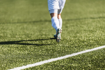 Back view of a soccer player running on a green grass pitch with white touchline. Close-up of legs in soccer cleats and white socks during a match or training. Concept of sports, motion