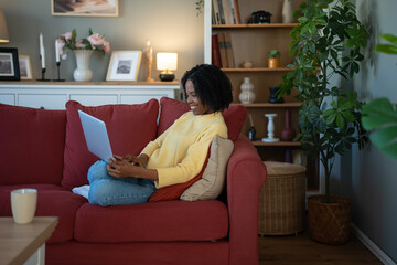 Obraz premium Young black woman smiling using laptop on sofa