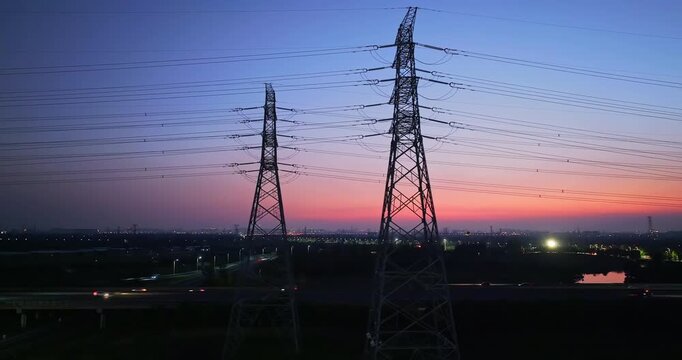 Electricity transmission pylons and high voltage power lines standing against beautiful sunset sky above city highway.