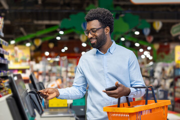 Adult man with eyeglasses smiling while holding an orange shopping basket and using a mobile device ,to make a contactless credit card payment at a self-checkout in a grocery store © Liubomir