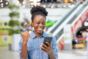 Happy young woman in a busy shopping mall celebrates good news on her smartphone, smiling and...