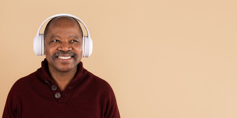 Smiling mature black man wearing white headphones listening to music on beige studio background....