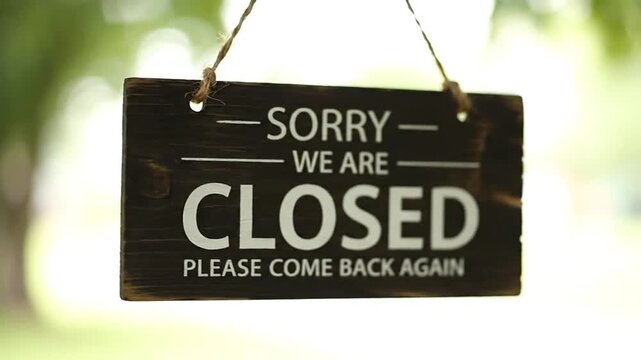 A black sign hanging from a rope, displaying a 'Closed' message, is visible against a blurred outdoor background with greenery and white light.