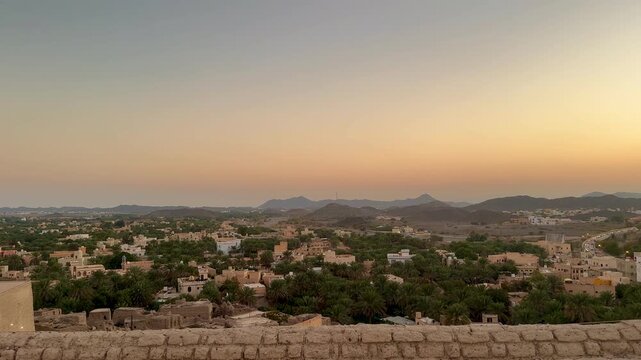 Panoramic view from Bahla Fort overlooking Bahla city with palm trees traditional houses mountains and local market under warm sunset sky. Bahla, Oman - February 22, 2026