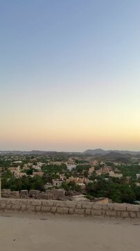 Panoramic view from Bahla Fort overlooking Bahla city with palm trees traditional houses mountains and local market under warm sunset sky. Bahla, Oman - February 22, 2026