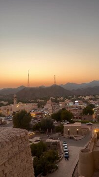 Panoramic view from Bahla Fort overlooking Bahla city with palm trees traditional houses mountains and local market under warm sunset sky. Bahla, Oman - February 22, 2026