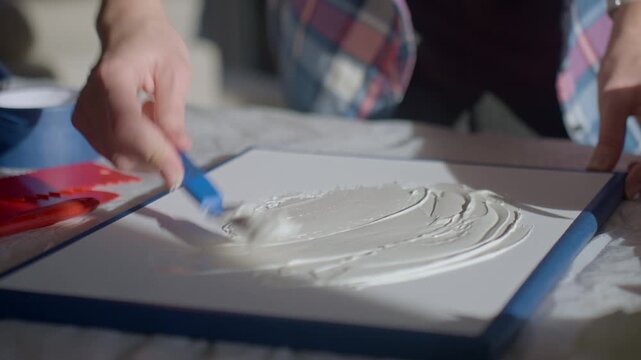 Close up of woman applying textured paste on white canvas using palette knife on work table