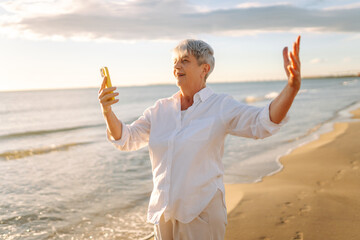 Gray-haired woman standing on beach, holding phone, talking on video call. Older woman takes selfie with sea in background, enjoying nature at sunset. Concepts: retirement, vacation, blogger.