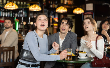 Naklejka premium Group of young sports fans upset and worrying during game match on TV in big beer pub