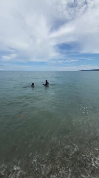 Two divers in wetsuits preparing to dive into the calm turquoise ocean water from a sandy beach on a bright, cloudy day.