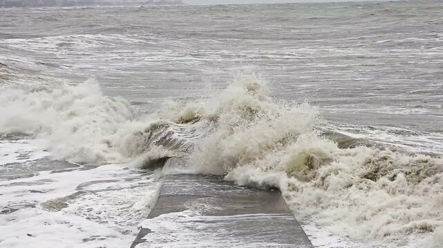 Stormy sea battering concrete jetty, relentless gray waves crashing over walkway, foamy surge and spray, winter gale driving erosion, dramatic coastal energy and storm surge motion