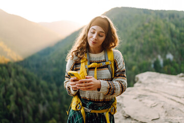 Beautiful woman with phone stands on rocky outcrop at sunset. Hiker with phone and backpack feels free and enjoys mountain scenery while hiking. Concept of nature, technology, and adventure. © maxbelchenko
