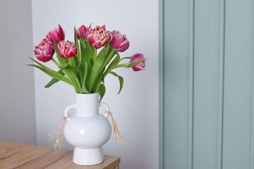 A beautiful bouquet of pink double tulips in a white ceramic vase stands on a wooden table against a sage green paneled wall. Concept of modern spring home decor. © Helga P-A.
