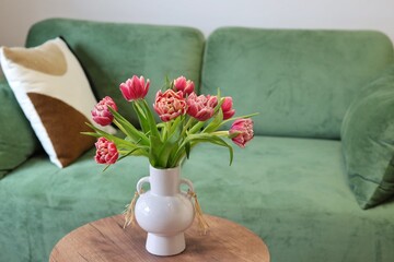 Fototapeta premium A bouquet of pink double tulips in a white ceramic vase on a wooden table in front of a green velvet sofa. Concept of cozy spring home decor. 