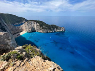 Fototapeta premium Aerial view of a stunning turquoise bay with a sandy beach surrounded by steep cliffs and lush greenery under a clear blue sky in Zakynthos, Greece
