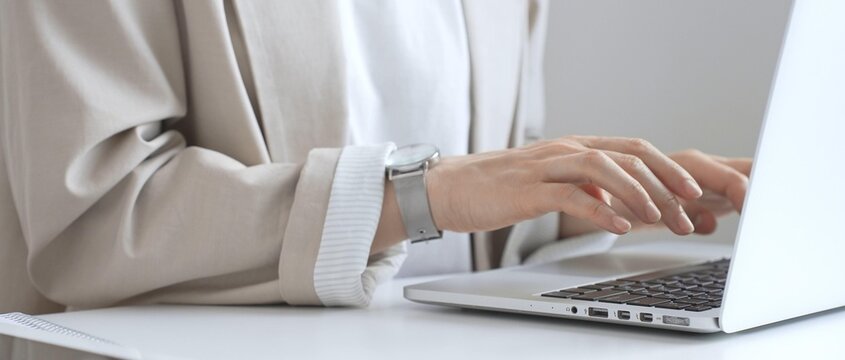 Professional businesswoman working intently on laptop, wearing tailored beige suit and silver watch, seated at minimalist white desk in modern office environment. Business people concept