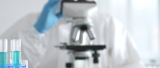 Fototapeta premium Male medical scientist in protective suit using microscope while analyzing samples, conducting research in laboratory. Close-up of test tubes with blue liquid