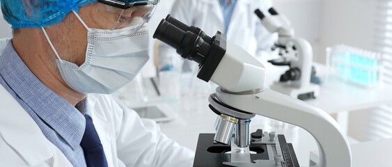 Senior male scientist wearing a protective mask, cap, and gloves, examining samples under a microscope in a modern laboratory. Medicine, healthcare and science concept