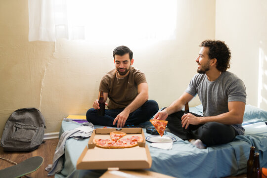 Young men enjoying pizza and beer in a casual room, roommates relaxing together in their basic apartment