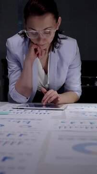 Businesswoman intently reviewing financial charts and reports scattered across her desk while using a digital tablet, working overtime in a corporate environment