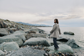 A young woman stands on rocky shore near calm sea under cloudy sky. She wears casual clothes and looks over her shoulder. Concept of nature, solitude, and outdoor lifestyle. © SHOTPRIME STUDIO