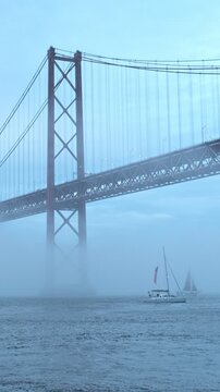 View of 25 de Abril Bridge famous tourist landmark of Lisbon connecting Lisboa and Almada in heavy fog mist with yacht boats passing under. Lisbon, Portugal