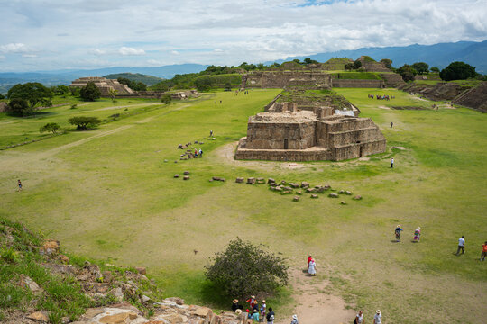Tourists explore ancient ruins under cloudy sky in Oaxaca, Mexico, visiting Monte Alban's stone structures surrounded by green grass and distant hills.