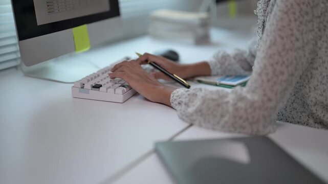 A woman is typing on a keyboard with a pen in her hand. She is sitting at a desk with a laptop and a book. The scene suggests a typical workday, with the woman focused on her tasks