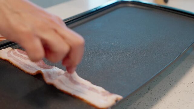 close up of hand placing raw bacon strips onto a hot sizzling electric griddle