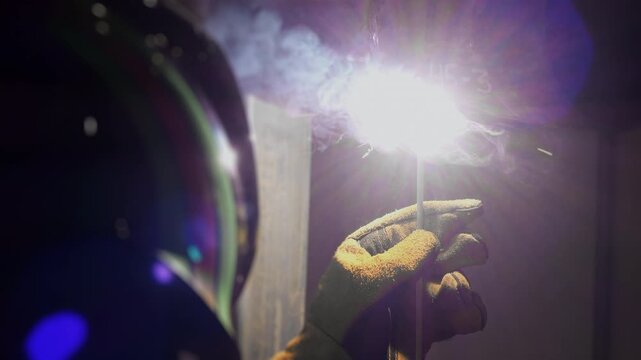 Close up of welder performing arc welding with electrode and bright electric arc while joining metal in industrial workshop.