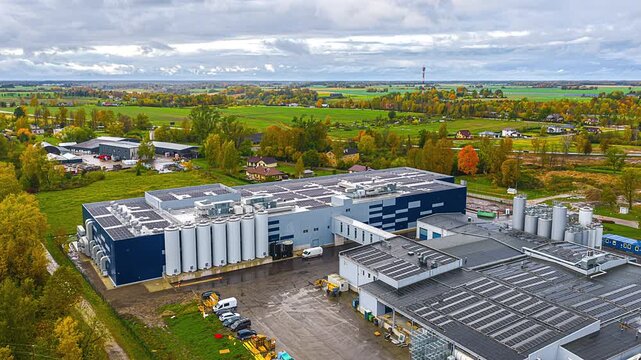 Drone hyperlapse over a large factory with rooftop solar panels and storage silos beside autumn farmland under cloudy sky, showing sustainable manufacturing and rural industry landscape.