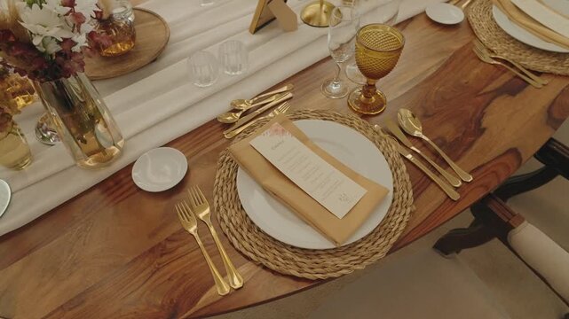 Overhead view of refined dining table arrangement with white plate woven placemat golden flatware menu card and floral decor