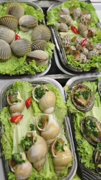 Vertical screen Fresh Sea Snails On Lettuce Trays, Vendor Arranges Glossy Shells With Chopped Green Onion And Chili, Closeup Texture Highlights Ridged Shells And Brine Sheen, Bustling Street