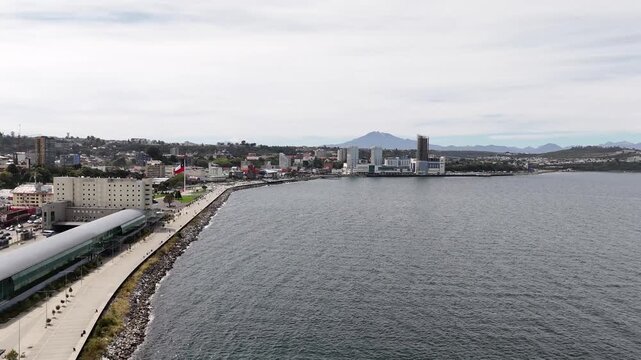 Cinematic aerial shot raising over the deep blue waters of Reloncav&iacute; Sound to reveal the Puerto Montt city skyline, the scenic Costanera waterfront, and the snow-capped Osorno Volcano on the horizon