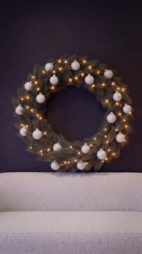 A beautifully decorated pine wreath with white ornaments and fairy lights hangs on a dark modern wall above a cozy white bed in a Scandinavian interior