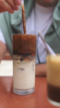 Vertical screen Person Drinks Cold Brew Outside. Individual Enjoying Chilled Layered Coffee At Sunny Outdoor Cafe. Man In Hoodie Savoring Multilayered Cold Coffee In Bright Street Cafe