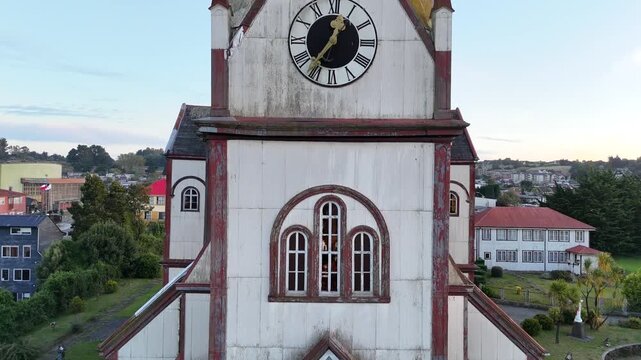 Aerial rises vertically, following the white corrugated iron facade and clock tower of the Sacred Heart Church (Iglesia del Sagrado Coraz&oacute;n de Jes&uacute;s), revealing the residential skyline of Puerto Varas