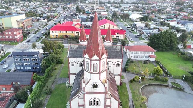 Aerial pulls back from the red spires of the Sacred Heart Church (Iglesia del Sagrado Coraz&oacute;n de Jes&uacute;s), revealing the full Romanesque-style structure and the surrounding urban grid of Puerto Varas