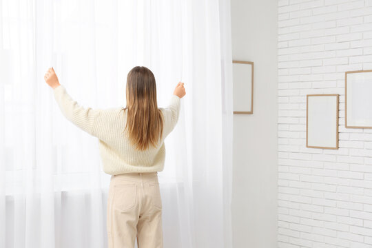 Woman closing curtains in light room at home, back view